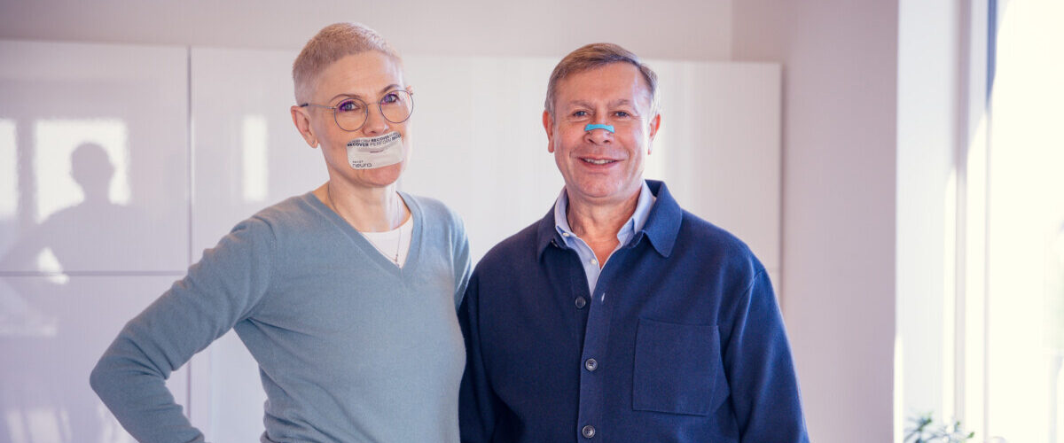 Two adults smile for the camera in a bright room; the woman has tape over her mouth and the man wears a blue nasal strip for a health-themed photo.