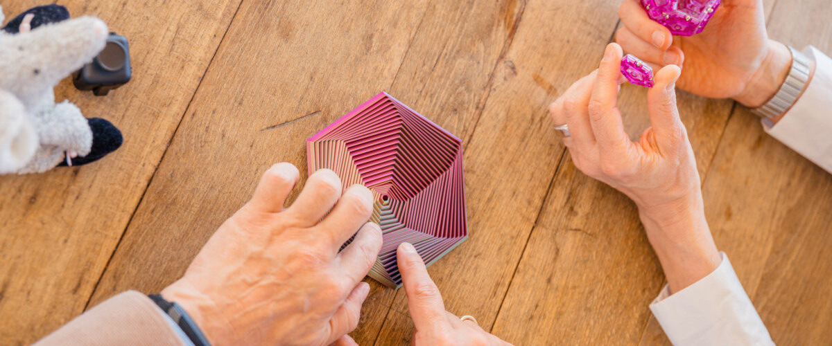 Hands of two people assembling a colorful modular origami sculpture on a wooden table.